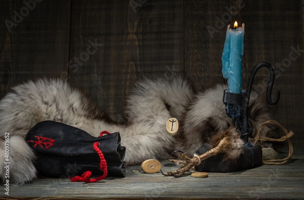 Fototapeta The Algiz rune is a rune of protection. A composition of runes, a dried bird's foot, a burning candle and an old leather bag for Scandinavian runes on the background of an animal skin. selective focus