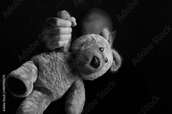 Obraz A man's hand holds a teddy bear by the paw, the focus is in the foreground. Conceptual photo: rude attitude towards children. Black and white photo.