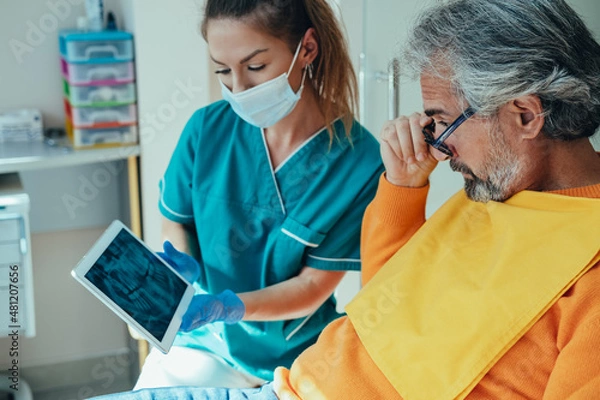Obraz Female Dentist Explaining Teeth X-ray to Surprised Mature Patient at Dentist's Office. 

Serious woman dentist talking to worried male patient and showing digital tablet with x ray film.
