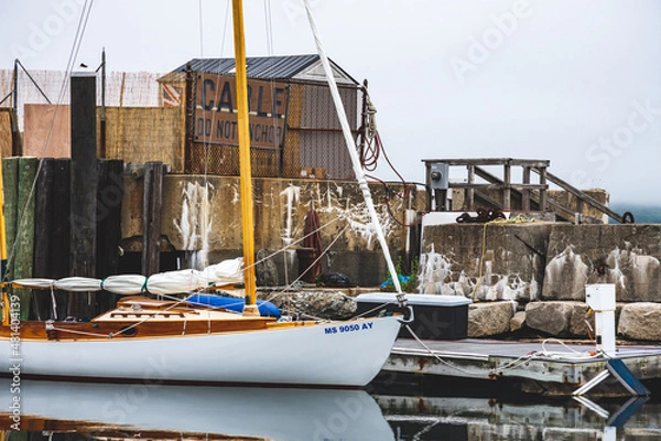 Fototapeta Sailboat docked near rustic shipyard on cloudy day in small seaside town