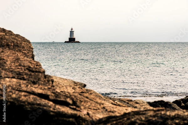 Fototapeta Lighthouse in ocean near rocky seaside on cloudy day in New England.