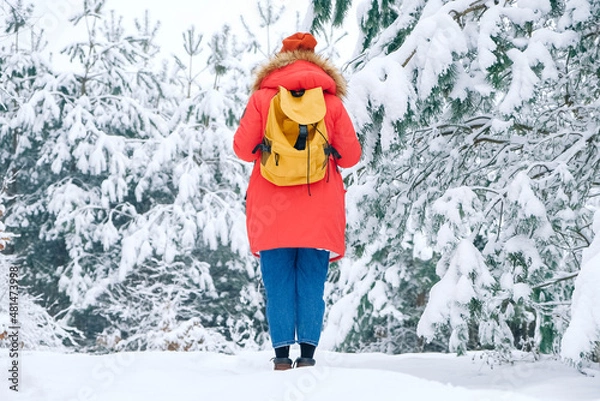 Fototapeta Woman in a red warm jacket and with a backpack on a background of snowy forest