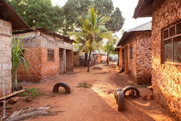 Fototapeta View on african village with palm. Street full of light, vibrant red soil and red color of buildings. Huts along the street. Traditional architecture, Tanzania countryside.
