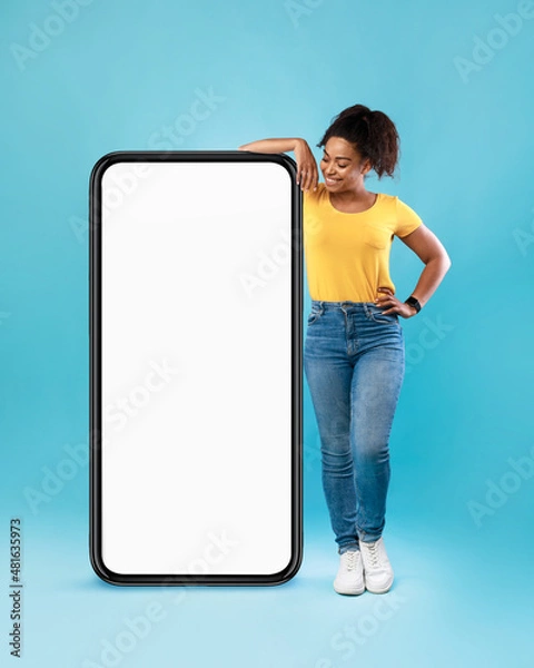 Fototapeta Joyful Afro woman in casual wear leaning on huge cellphone with empty white screen on blue background, mockup