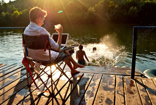 Fototapeta A man with a laptop and a glass of alcohol sitting in a chair. The children relax on the pier and jump into the water. The concept of summer, recreation, responsibility and supervision.