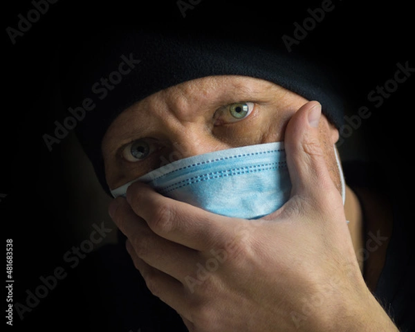 Obraz A man in a protective mask covered his mouth with his hand, close-up. Viral infection Covid-19 (SARS-CoV-2). Respiratory protection. Pandemic and deadly Omicron virus.

