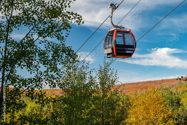 Obraz Cable car on a cable passes by nearby trees at a ski hill in Minnesota, with autumn colors on the distant hill and mostly blue sky.