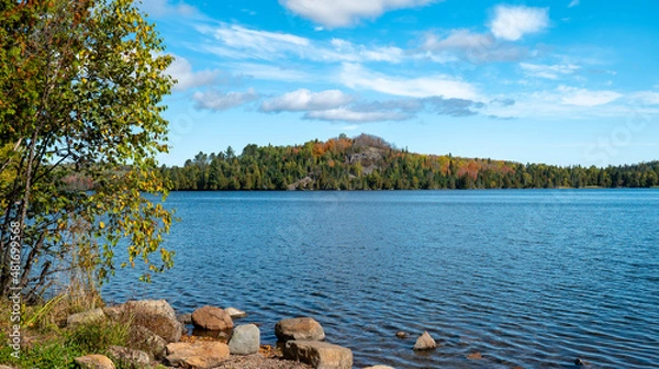 Obraz Beautiful blue water and sky at a lake in northern Minnesota on a sunny autumn day, with boulders along near shore and trees on rocky hill on the distant shoreline.
