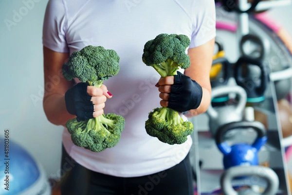 Fototapeta Woman in the gym lifts a broccoli in the form of a dumbbell