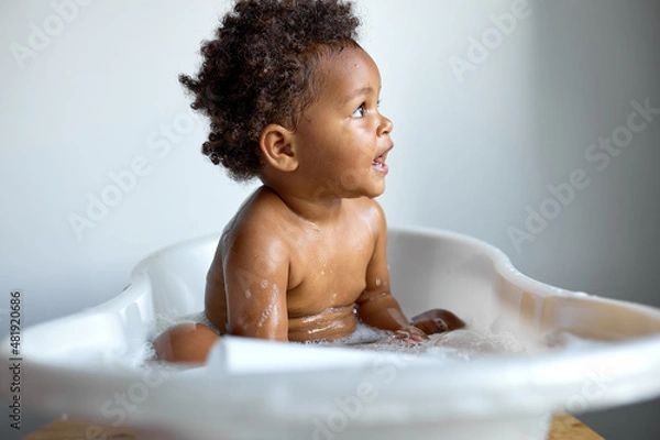 Fototapeta Small cute black child sitting at bath, washing, playing with soap and foam, indoors, copy space, white background, happy childhood, banner. adorable kid enjoying hygiene skincare procedures