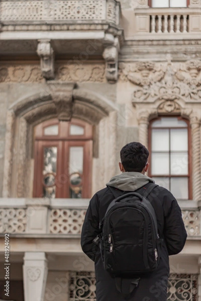Fototapeta Back pose of young tourist boy wearing mask. He has back pack. He is looking to historical old building. The focus is on boy. Background is flu. Vertical photo.
