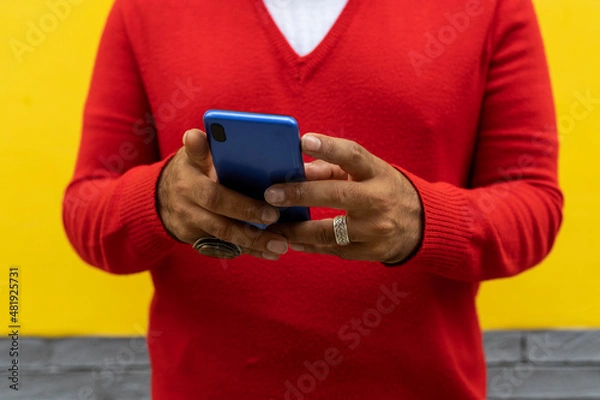 Fototapeta Adult male holding a blue cell phone with both hands. He is wearing a red sweater and white flannel. Yellow and gray wall background. Technology concept.