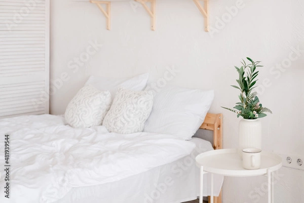 Fototapeta White cozy bedroom with a green plant on the table. Empty bed with a white bedding set,