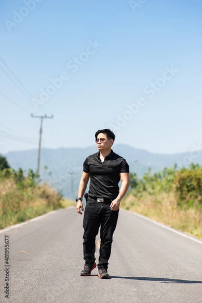 Obraz thai asian man short hair serious face wear black shirt plat wrist watch and shade walking with confident bollywood style on middle of asphalt street and mountain in background