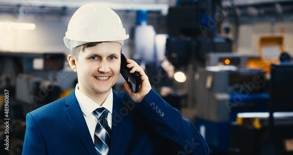 Fototapeta Portrait of a professional heavy industry engineer in protective uniform and hard hat, smiling charmingly. Unfocused large industrial plant in the background