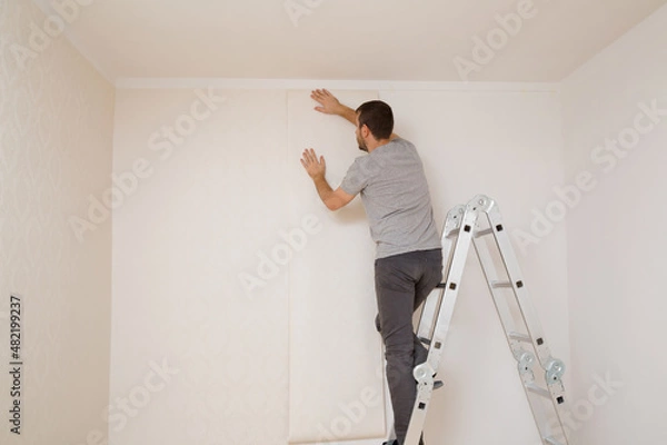 Fototapeta Young adult man standing on metal ladder and applying new wallpaper on white wall in room. Repair work of home. Making interior change. Back view.