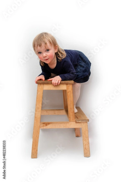 Fototapeta A two-year-old girl in a dark blue dress climbs onto a two-step chair. He stands on the first step and looks into the camera. Blonde with blue eyes. On a white background.