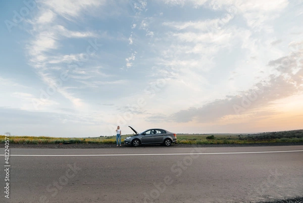 Fototapeta A young girl stands near a broken-down car in the middle of the highway during sunset and tries to call for help on the phone. Waiting for help. Car service. Car breakdown on road.