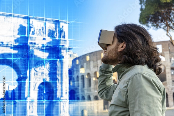 Fototapeta Young man in Piazza del Colosseo (Rome) looks at a virtual reconstruction with a cardboard viewer. The landscape around him changes, taking him on a journey through time and space.