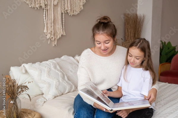 Obraz mother and daughter are sitting at home on the bed flipping through and looking at a book with photos. important moments of life in a photo album. Cute sisters read an interesting fairy tale together
