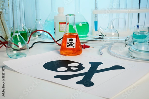 Fototapeta Death printed symbol on a chemical table. Poison fluids in toxicology laboratory. Photo contains danger paper sheet, test tubes, orange colored liquid in chemical flask, some retorts, electrodes
