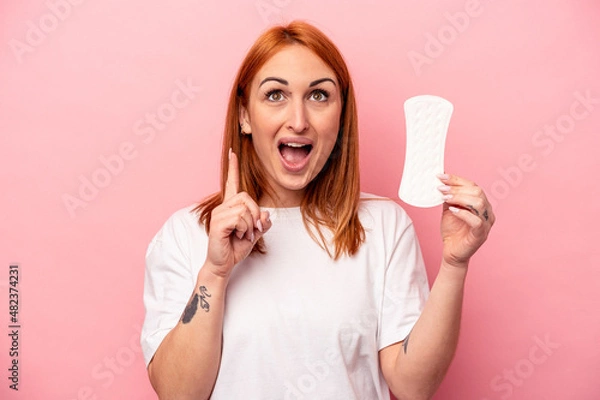 Obraz Young caucasian woman holding sanitary napkin isolated on pink background pointing upside with opened mouth.