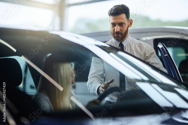 Fototapeta Kaukaski sprzedawca pokazujący nowemu luksusowi samochodowi klienta w salonie. Szczęśliwa młoda kobieta wybierająca nowy pojazd w Auto Salon. Zakup i koncepcja ludzi.