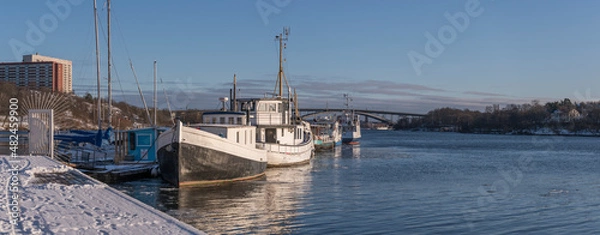 Fototapeta Harbor view with boats at a jetty on the island Lilla Essingen with skyline of downtown a sunny and snowy winter day in Stockholm