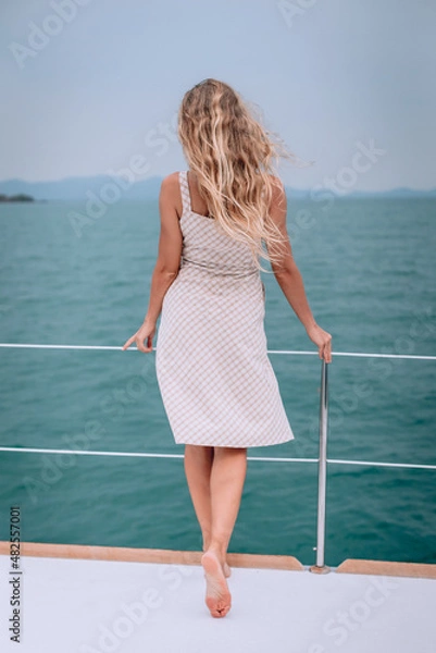 Fototapeta Summer photo from a vacation to the sea from a yacht. Feminine and elegant image with a slim girl with long curly hair turned back in a beautiful plaid summer dress. The observation of the landscape.