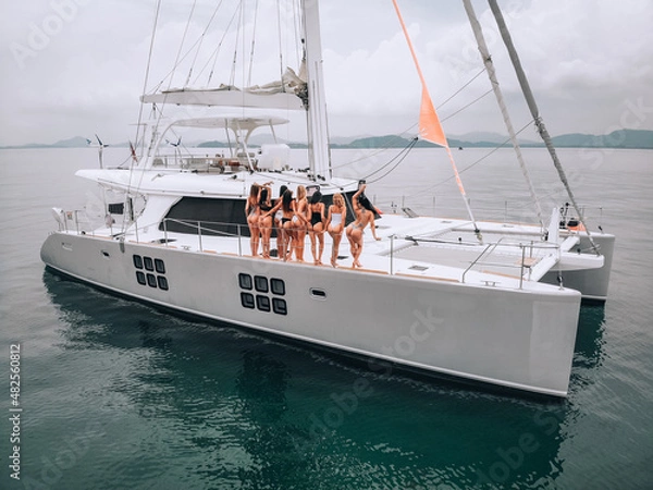 Fototapeta Good looking young girls in different swimsuits stand on a white yacht and pose for the camera in the sky. Tropical background of green islands. Drone photo session. The concept of travelling.
