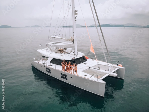 Fototapeta A photo created by drone. A lovely young women in a beautiful swimsuit pose on a white expensive luxury yacht in the bay open sea.Demonstration of luxury life. The concept of women's beauty.