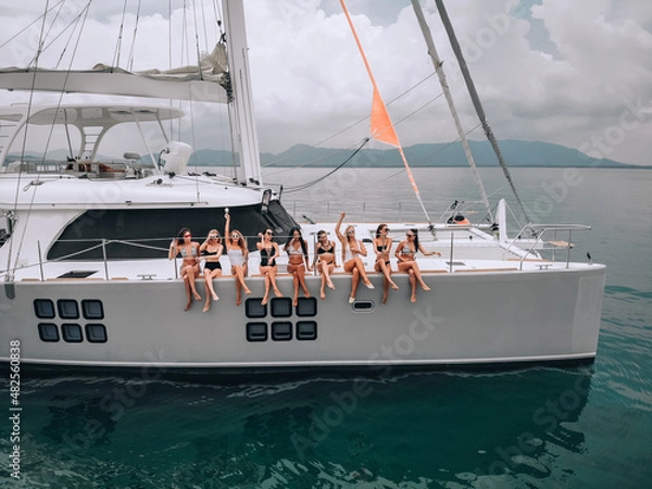 Fototapeta Photo from a drone with young cute girls in swimsuits and sunglasses posing sitting on a luxury white yacht in the open sea. The background is the shore. The concept of a holiday photo session.