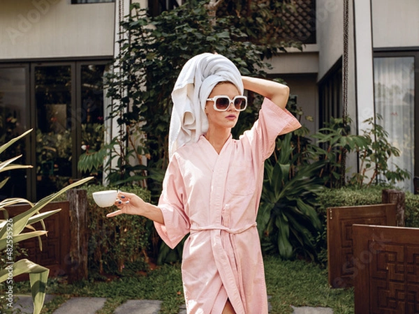 Fototapeta Portrait of a sexy and calm young attractive model in a bathrobe, towel and sunglasses posing against the backdrop of greenery and a villa. Morning concept