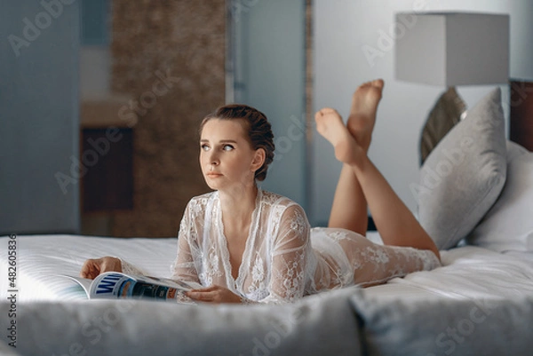 Fototapeta Attractive innocent cute girl in a white lace transparent nightie lying on a snow-white bed at home and holding a magazine in her hands. Comfort concept