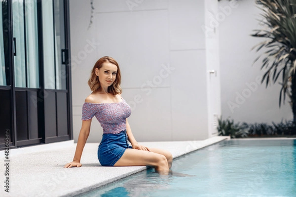 Fototapeta Cute young slender model wearing a colored jacket and shorts is sitting by the pool against the background of a bright white house. Holiday concept