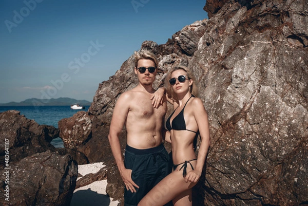 Fototapeta Portrait of a beautiful young couple man and woman wearing swimwear and posing next to a rock, looking straight into the camera with their heads held high