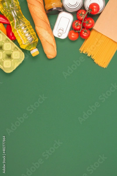 Obraz Vertical image with groceries, food donations on green background with copy space - pasta, vegatables, canned food, baguette, cooking oil, tomatoes. Food bank, food delivery concept