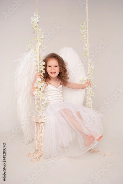 Obraz a 5-year-old girl rides a swing in an angel costume in a beautiful white dress and wings for Valentine's Day February 14 in love