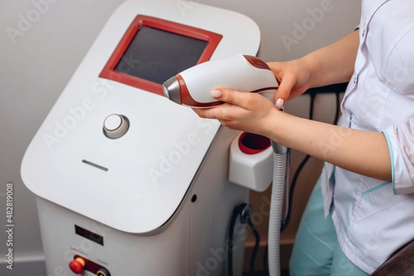 Fototapeta A white medical device standing in a beauty salon. A woman in uniform holds in his hands ascentic to get to work. Working with a facial skin treatment device. The concept of health.
