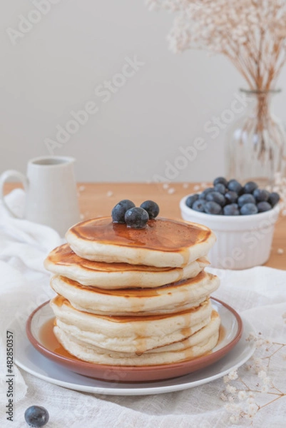 Fototapeta Vertical image of a stack of pancakes with blueberries on top and maple syrup or honey running down the sides. Fruit, flowers and jug on top a wooden table. Copy space available in light airy backdrop