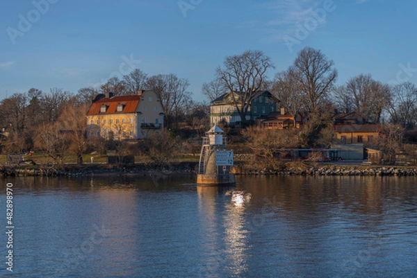 Fototapeta Panorama view over old houses and a lighthouse with a speed sign by the water front at the ness Blockhusudden a sunny winter afternoon in Stockholm 