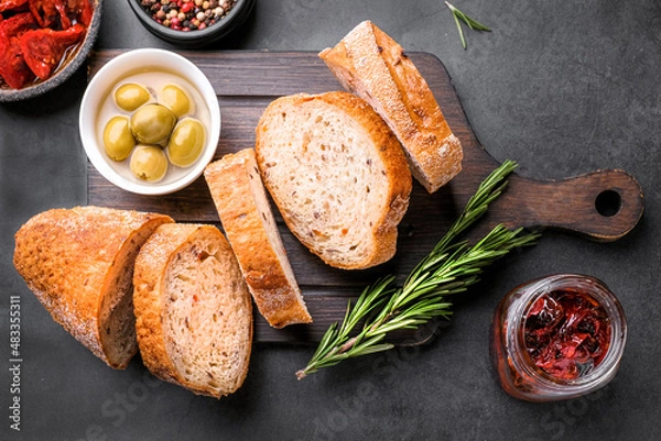 Fototapeta Slices of Ciabatta bread  on wooden board with olives, dried tomatoes, oil, rosemary and herbs on rustic wooden background. top view.
