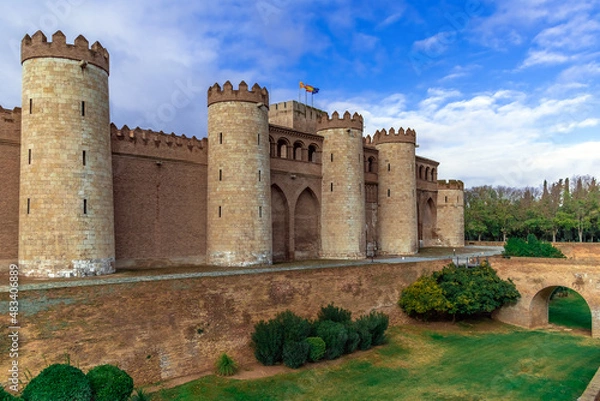 Fototapeta Zaragoza, Spain - November 29, 2021: Medieval stone facade with towers of Aljaferia Palace in Saragossa - outside view. Ancient fortress, moat around with green grass and bushes and arch bridge