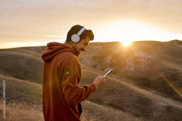 Obraz fit caucasian happy man in headphones chatting with someone on smartphone, typing message and smiling outdoors, having rest, enjoy time at sunrise or sunset. in mountains in nature