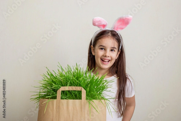 Fototapeta A cute little girl with pink rabbit ears stands next to a paper bag with grass