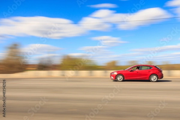 Fototapeta A red car is driving fast on the road on a sunny summer day, the car is in focus, the background is blurred.