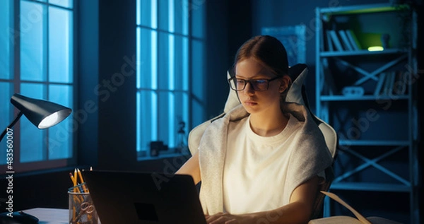 Fototapeta Intelligent caucasian young girl teenager uses computer to watch online school lessons, doing homework. Bright college student studying from home, preparing for exam till late at night.