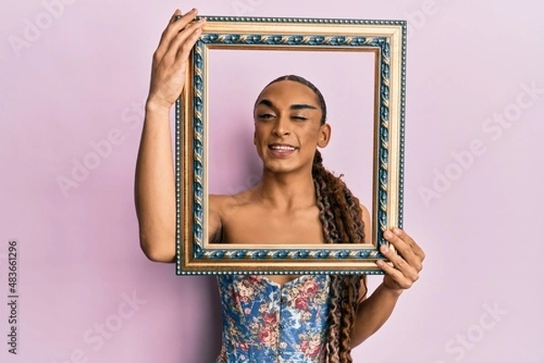 Fototapeta Hispanic man wearing make up and long hair holding vintage empty frame winking looking at the camera with sexy expression, cheerful and happy face.