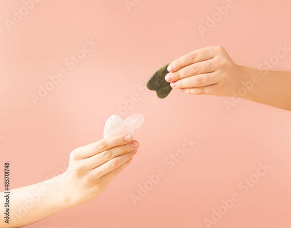Fototapeta Women's model hands with a scraper made of green and pink jade gua sha on a beige background. Spa means for body care. Gua sha stone scraper for beauty care. Copy space