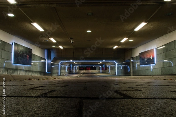 Fototapeta A tunnel underground for bikers and pedestrians, bikers in the back riding their bikes and having their lights on. The lights in the tunnel reflection in the walls